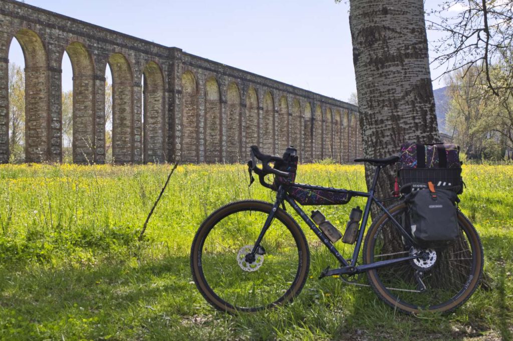 Ein Gravelbike mit Fahrradgepäck ist an einen Baum gelehnt. Dominant im Hintergrund befindet sich das Acquedotto Del Nottolini, das Aquädukt von Lucca in der Toskana.