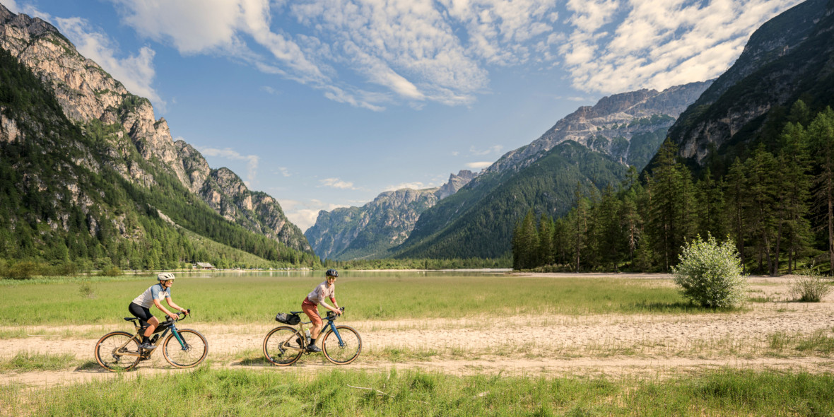 Zwei Gravelbikerinnen fahren einen schönen Schotterweg entlang, im Hintergrund sieht man die Berge der Alpen