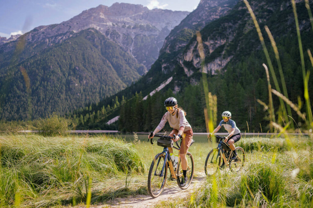 Zwei Fahrradfahrerinnen fahren mit dem Gravelbike auf einem unbefestigten Weg an einem See in den Dolomiten.